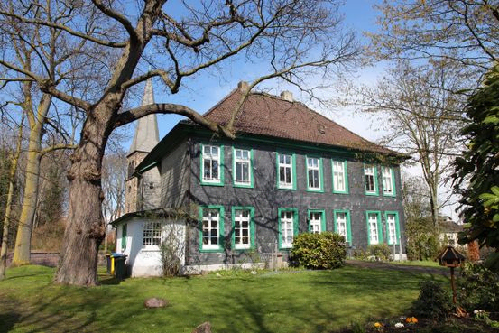 Aufnahme des alten Pfarrhauses an einem sonnigen Tag. Das Gebäude wird vollständig abgebildet. Links im Hintergrund ist der Kirchturm zu sehen.